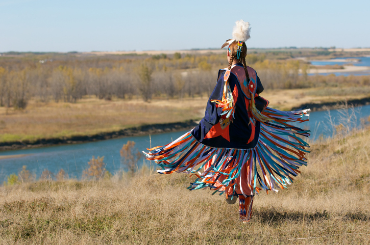 A person in American Indian clothes on empty land, beside a water body