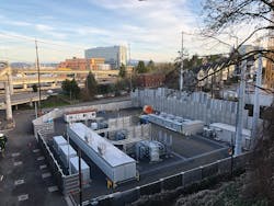 This view of the completed Marquam substation shows the 115-kV, four-bay, breaker-and-a-half gas-insulated substation in the background (left) next to the relaying control enclosure (right), located in front of the retaining wall. In the foreground (right to left) are three 50-MVA transformers, a 39-unit networked switchgear and three 13-kV metal-enclosed shunt capacitor banks. On the far left, next to the relaying control enclosure, four vaults collect the distribution circuits and route through the underground bores into downtown Portland.