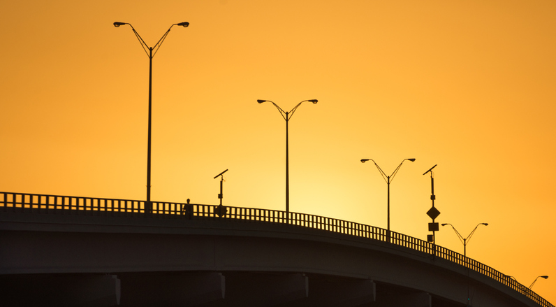 A man is walking over a bridge at sundown that spans the Indian River in Vero Beach, Florida. With the sun setting behind the bridge, the whole bridge is bathed in golden light.