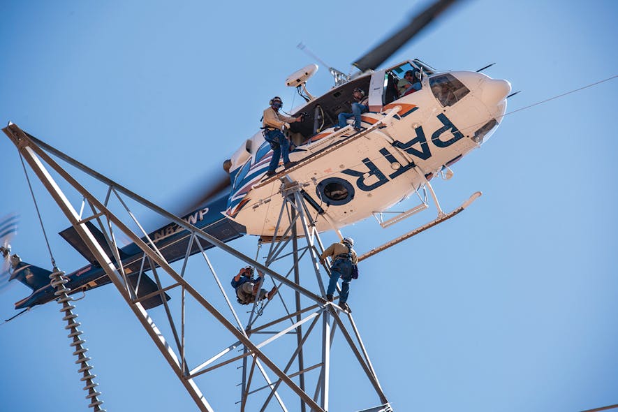 Aerial line workers descend from aircraft onto transmission tower to conduct inspection.