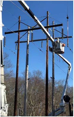 A Three Phase Line Construction lineman in bucket (foreground) wears his bare-hand live line suit and hood to transfer an energized conductor from an old, wooden H-frame tower to a steel H-frame. A Three Phase Line Construction lineman in bucket (foreground) wears his bare-hand live line suit and hood to transfer an energized conductor from an old, wooden H-frame tower to a steel H-frame.