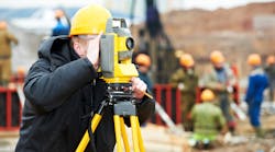 A surveyor peers through a theodolite at an outdoor construction site. Underground obstacles must be considered when burying power lines.